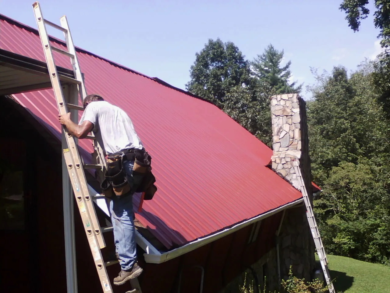 Roofer on a ladder beside a red metal roof and stone chimney — Soffit & Fascia Repair by Roofing Brothers in Antrim